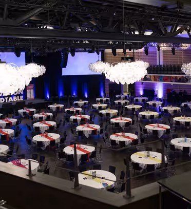 Spacious conference room set for an event, featuring numerous round tables with white tablecloths and colorful runners. A large stage with blue lighting and a screen is seen at the front, and elegant chandeliers hang from the ceiling.