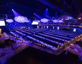 A large event hall is set for a formal gathering. Long banquet tables are adorned with table settings and surrounded by chairs. Blue and purple lighting creates an elegant atmosphere, with decorative chandeliers hanging from the ceiling.