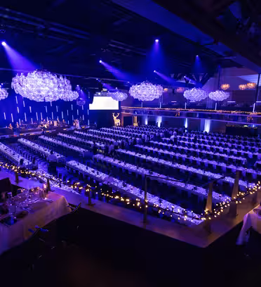 A large event hall is set for a formal gathering. Long banquet tables are adorned with table settings and surrounded by chairs. Blue and purple lighting creates an elegant atmosphere, with decorative chandeliers hanging from the ceiling.
