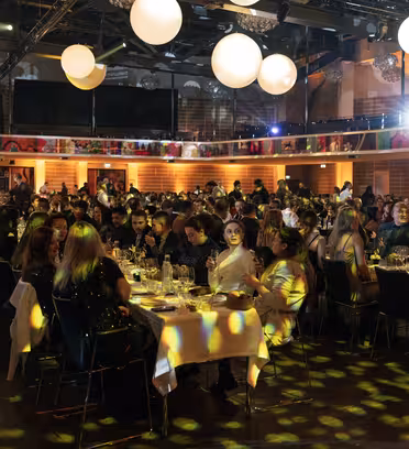 A large event hall filled with people seated at tables, socializing and dining. The room is adorned with large, glowing spherical lights and colorful decorations. The atmosphere is lively, with attendees enjoying food and conversation.