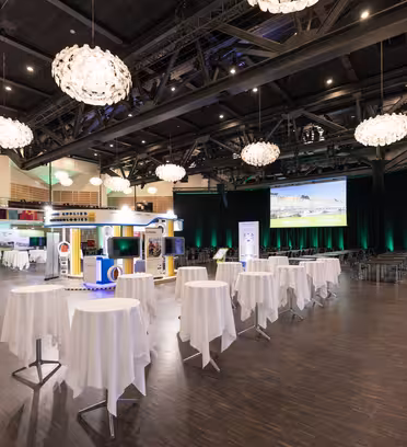 Spacious event hall with round tables covered in white tablecloths, large screen displaying visuals, attendees walking, and green lighting accents. Ceiling features large, ornate chandeliers. Exhibition booths line the walls with various displays.