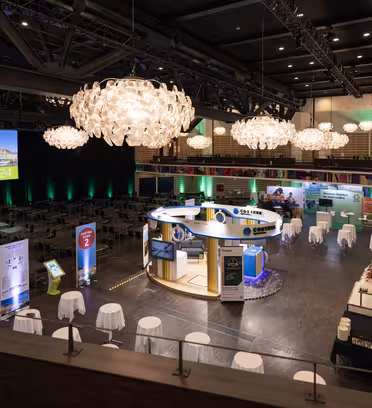 A spacious convention center is set up for an event, featuring multiple exhibit booths, signage, and round tables with white tablecloths. Large chandeliers hang from the ceiling, and a screen displays Welcome 2024.