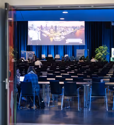 View through open doors of a conference room with rows of empty chairs facing a stage. A large screen displays a cityscape image next to a NAME CHANGE banner. Blue curtains and potted plants decorate the space. A few people are seated at tables.