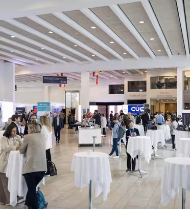A brightly lit conference room with groups of people networking around white round tables. Banners and signs are visible in the background and the room has a modern design with high ceilings and natural light.