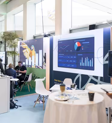 A modern conference room with information screens displaying charts and graphs. People are engaged in discussions around a booth with brand displays and conference materials. The atmosphere is professional and business-oriented.