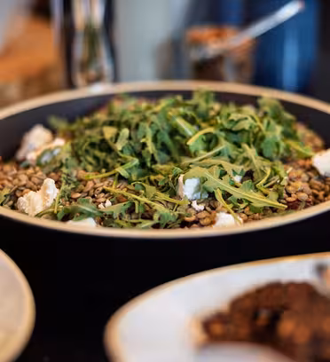 A close-up of a black bowl filled with lentils, arugula, and chunks of cheese. The background shows other dishes partially visible on a dark tabletop.