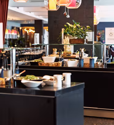 Buffet setup in a restaurant with various salads in white bowls on dark counters. Colorful lamps hang from the ceiling, and potted plants add greenery. In the background, tables with purple tablecloths and curtains are visible.
