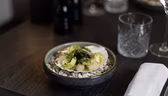 A dish with scallops, avocado, and herbs is elegantly plated on a bed of rocks in a dark bowl, placed on a wooden table. Nearby are a glass tumbler, a white napkin, and blurred background items including a pepper grinder and a lamp.