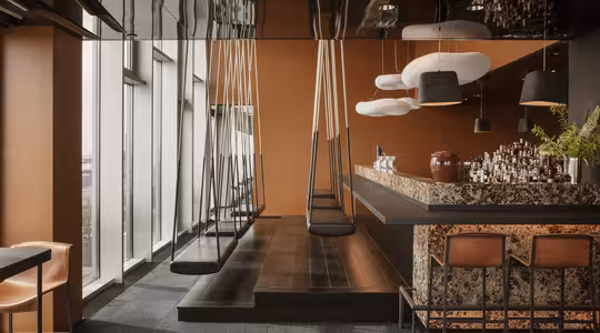 A modern bar setting featuring a counter with high chairs, glass jars, and hanging lights. Swings are used as seating at a sleek, dark wood communal table. The room has large windows and a warm, brown color palette.