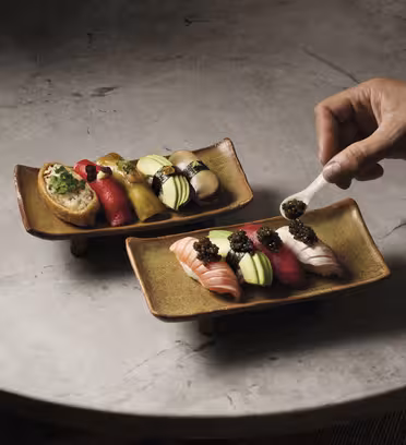A person placing caviar on sushi with a spoon. Two plates of sushi are on a stone surface, featuring variety like salmon, tuna, avocado, and garnish. The sushi pieces are artfully arranged in vibrant colors.