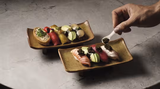 A person placing caviar on sushi with a spoon. Two plates of sushi are on a stone surface, featuring variety like salmon, tuna, avocado, and garnish. The sushi pieces are artfully arranged in vibrant colors.