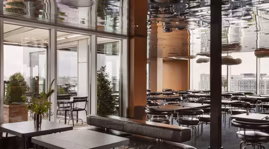 Modern dining area with sleek black tables and chairs, large glass doors leading to a balcony, and a reflective ceiling. Natural light illuminates the space, highlighting green plants and an urban view in the background.