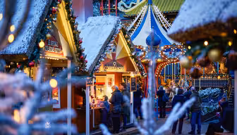 A festive Christmas market scene at dusk, featuring small wooden stalls adorned with lights and decorations. People gather around a coffee stand, and snow-dusted trees frame the colorful, illuminated surroundings.