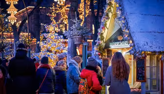 People gather around a festive market stall adorned with lights and snow, in a winter setting. Warm clothing, including coats and scarves, suggest the cold weather. In the background, more lights decorate trees, creating a cozy, festive atmosphere.