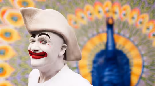 A person dressed as a clown with white face paint, red lips, and a large white hat smiles and winks. They stand in front of a colorful peacock mural, enhancing the vibrant background with eye-like patterns.
