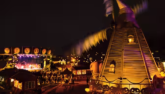 A vibrant nighttime Halloween scene features a lit-up windmill and festive decorations, including pumpkins with carved faces. A stage in the background hosts a performance, and people are gathered around, enjoying the illuminated atmosphere.
