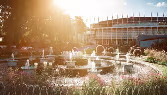 A serene garden scene with a round fountain in the foreground, surrounded by flowers and plants. A tall building with numerous flags on top is in the background. The sun casts a warm glow across the image.