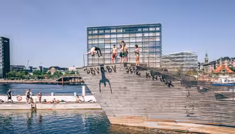 People dive and sit on a wooden ramp by the water, with modern buildings in the background. The sunny day shows a lively, recreational scene by the waters edge.