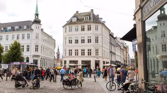 A bustling European city square on a cloudy day, featuring cobblestone streets, people walking, and some with strollers and bicycles. The area is surrounded by historic buildings with a café visible on the right.