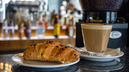 A flaky croissant on a white plate beside a frothy cappuccino in a glass, set on a dark countertop. The background shows a blurred coffee machine and various bottles, suggesting a cozy café setting.