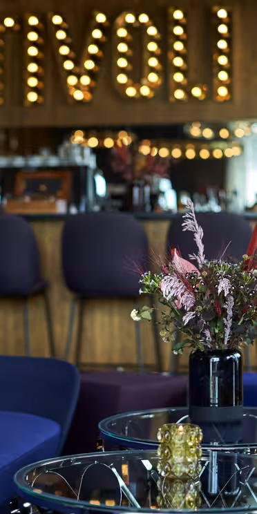A stylish lounge area with blue and yellow seating around a glass table topped with flowers. A bar with high stools is in the background, and large lightbulb letters spell TIVOLI on the wall.