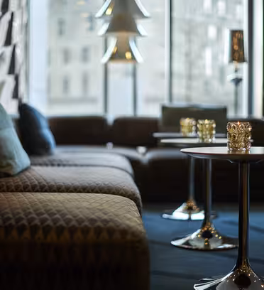 A modern lounge area with quilted sofas, small round tables, and decorative lamps. Soft natural light filters through large windows, highlighting the chic decor and creating a cozy atmosphere.