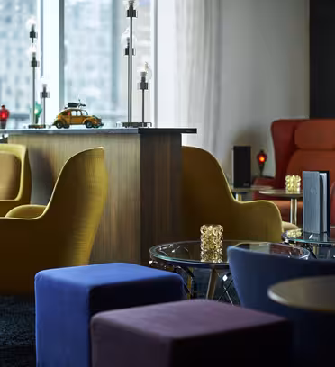 A modern lounge area featuring colorful chairs in yellow, red, and blue. Glass tables with decorative lamps and a wooden counter are visible. Large windows allow natural light to brighten the room, with an urban skyline in the background.