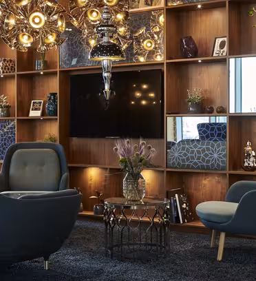 A modern living room with a cozy seating area featuring blue armchairs and a round glass coffee table. A wooden bookshelf with decorative items and a TV is in the background. Unique pendant lights and a floor lamp add ambiance.