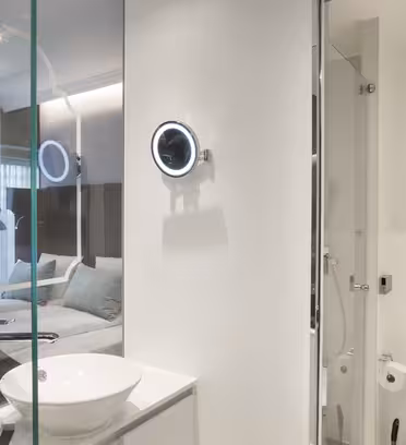 Modern hotel room with a glass-walled bathroom. Features include a white sink, illuminated mirror, and toilet. Partial view of a bedroom with a bed and a blue armchair visible through the glass. Soft lighting creates a sleek, contemporary atmosphere.