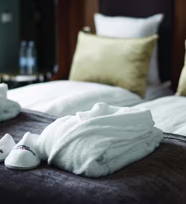 Two neatly folded white bathrobes and a pair of white slippers are placed on a luxurious brown bedspread. In the background, a bed with white pillows and gold cushions is visible. Two water bottles are on a side table.