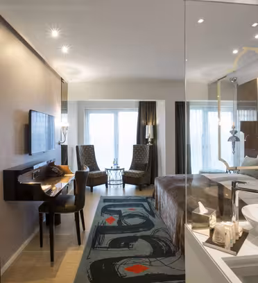 Modern hotel room with a large bed, flat-screen TV, desk, and two armchairs facing a window. The room features warm lighting, wooden flooring, and a unique abstract rug. The bathroom area is partially visible with a sink and mirror.