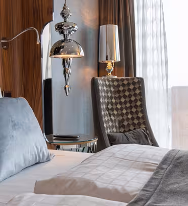 A modern hotel room with a neatly made bed featuring white and gray bedding and a gray pillow. A stylish lamp hangs beside a textured wall. A checkered chair is near a window showing a view of city buildings outside.