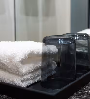 A bathroom countertop with two stacked white towels and two upside-down transparent drinking glasses on a black tray. A mirror reflects part of the scene. The countertop has a dark, marbled surface.