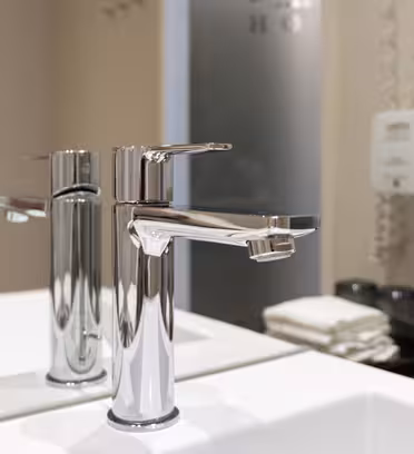 A modern bathroom sink with a chrome faucet is shown. The reflection of the faucet is visible in the mirror behind. To the right, theres a stack of folded towels and wall-mounted soap dispensers. The setting is clean and minimalist.