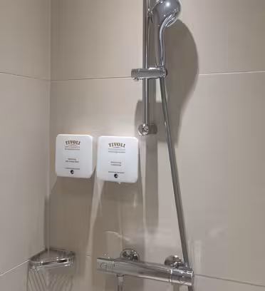 A modern shower with a chrome adjustable showerhead on a beige tiled wall. Two white wall-mounted soap dispensers labeled Tivoli are positioned below the showerhead. A small metal basket is attached to the wall.