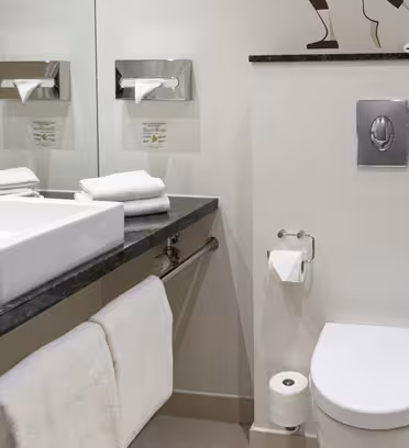 A modern bathroom with a white square sink on a dark countertop, chrome faucet, and a large mirror. Towels are neatly folded on the counter and bar. A toilet is nearby, and theres minimal decor with light walls and a clean design.