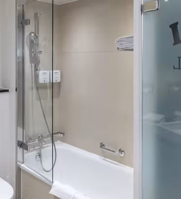 Modern bathroom with a bathtub-shower combo, glass partition, and wall-mounted shampoos. Toilet is next to the tub, and a towel is on the rack. Frosted glass door with partial text. Wall art of a cartoon figure on the left.