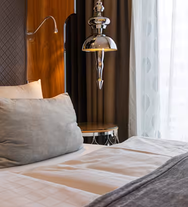 A neatly made bed with white linens and a gray pillow is beside a wooden panel with a silver lamp. Sheer curtains allow soft light to enter the room, creating a tranquil ambiance.
