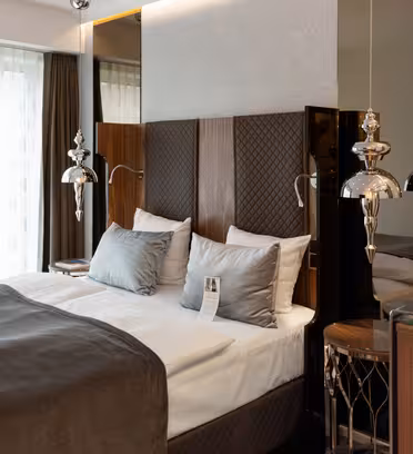 Modern hotel room with a double bed featuring gray sheets and pillows. A pamphlet rests on the bed. Two stylish metallic lamps hang above nightstands, with a large window covered by sheer curtains in the background.