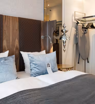 Hotel room interior with a neatly made double bed. Features blue and white pillows, a dark gray blanket, and wooden headboard. An ironing board and iron are in the open closet. A modern lamp hangs from the ceiling, adding a stylish touch.