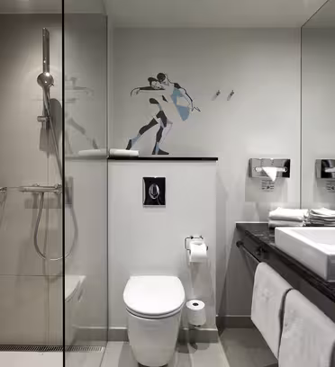 Modern bathroom with a glass shower enclosure, wall-mounted toilet, and large mirror. Double sinks with chrome faucets and toiletries on the counter. Wall art of a dancing couple is above the toilet. Towel hangs below the counter.