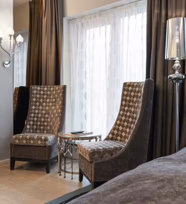 A modern hotel room features two patterned armchairs, a small glass-top table, and a tall mirror lamp beside floor-to-ceiling windows with sheer curtains. A flat-screen TV is mounted on the wall above a small desk with a black chair.