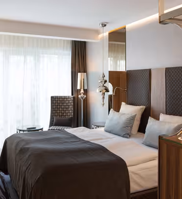 Hotel room with a double bed featuring white linens and gray blankets. A patterned chair and small table sit near large windows with sheer curtains. A desk with a chair and a few decorative elements add to the modern, elegant décor.
