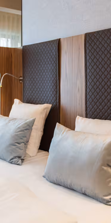A modern hotel room with a neatly made double bed featuring white and silver-gray pillows. The room has elegant decor, including quilted headboards, stylish pendant lights, a patterned armchair, and a side table by the window with sheer curtains.