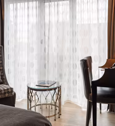 A hotel room with a patterned armchair, glass-topped table, and a black desk with a phone. Sheer curtains on large windows allow light to fill the space, while dark brown drapes frame the scene. A section of a bed is visible in the foreground.