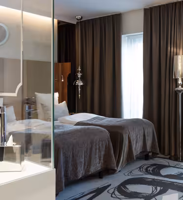 Modern hotel room with two twin beds covered in gray blankets. There’s a glass-walled shower area with a white basin sink. Brown curtains cover the windows, and a patterned chair is in the corner. Contemporary decor features metallic accents and abstract art.