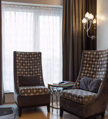 Two patterned armchairs with cushions face a round glass-topped table by a window with sheer curtains. A floor lamp and a wall-mounted TV are on the side, complemented by dark drapes and a wall sconce light fixture.