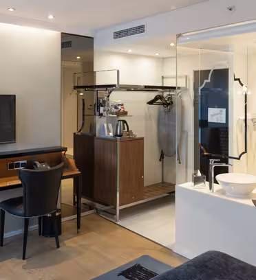Modern hotel room with a sleek design, featuring a wall-mounted TV, a desk with a chair, a glass-walled bathroom, and an open wardrobe with a hanging rail. Neutral color palette with wood flooring and contemporary lighting.