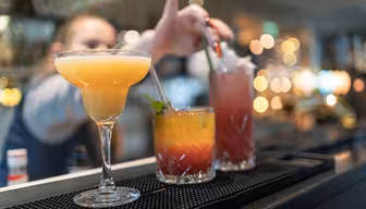 A lineup of colorful cocktails on a bar counter: a yellow margarita, a layered orange-red drink with mint and a straw, and a tall pink drink with a straw. A bartender adjusts a straw in the background, with blurred lights creating a warm ambiance.