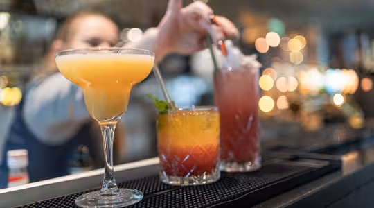 A lineup of colorful cocktails on a bar counter: a yellow margarita, a layered orange-red drink with mint and a straw, and a tall pink drink with a straw. A bartender adjusts a straw in the background, with blurred lights creating a warm ambiance.